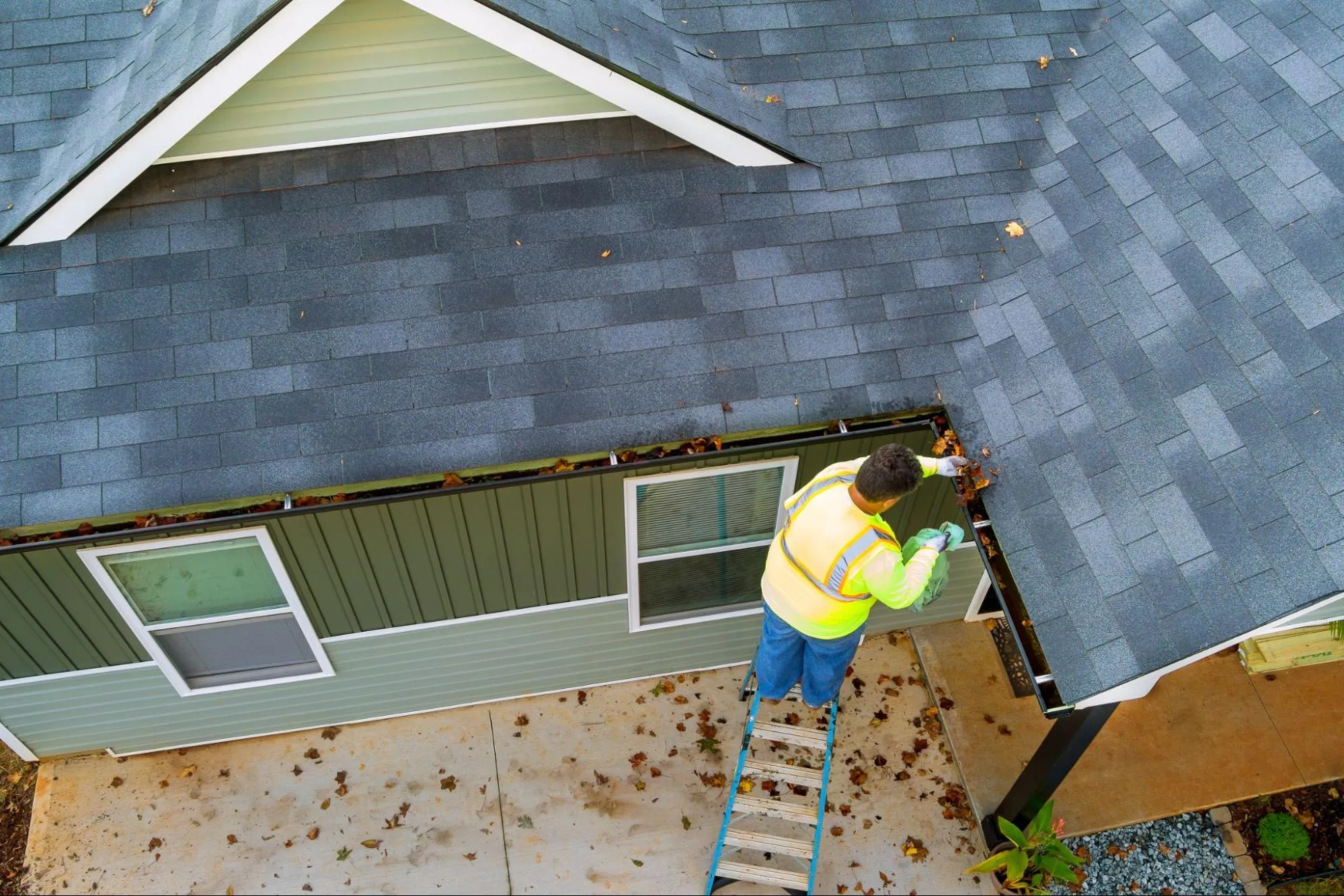A worker is on a ladder, cleaning a roof gutter.